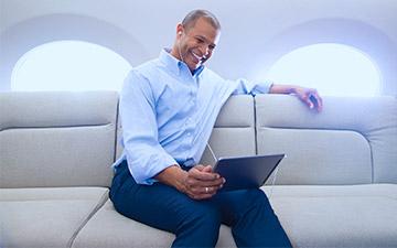 微笑的男人坐在私人飞机上，用私人飞机上网, 穿着蓝色纽扣衬衫看着平板电脑