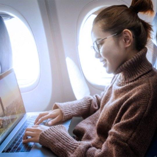一位女士戴着眼镜，穿着棕色毛衣，在飞机上通过无线网络用笔记本电脑工作