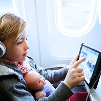 一个小女孩戴着耳机，腿上放着一个洋娃娃，在飞机上通过无线网络玩平板电脑游戏
