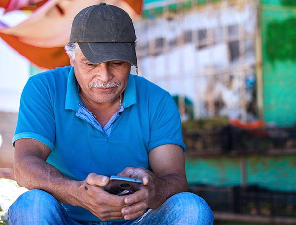 一名戴着帽子、穿着蓝色马球衫的男子，坐在一家商店外面看他的智能手机