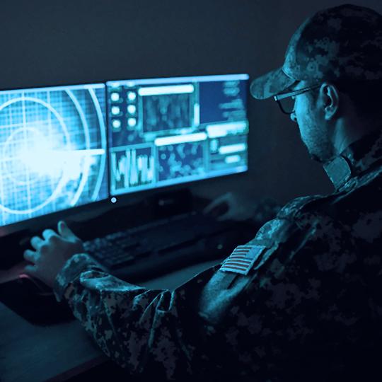 Man in camo uniform and cap wearing glasses sittng in the dark looking at two computer monitors